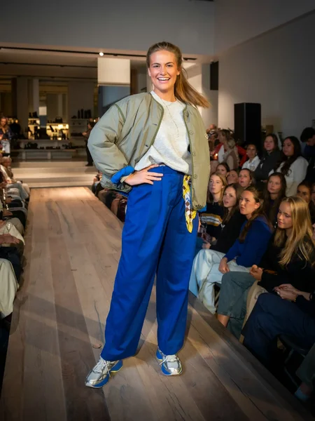 Groene bomberjas met grijze sweater, blauwe broek en kleurrijke sneakers
